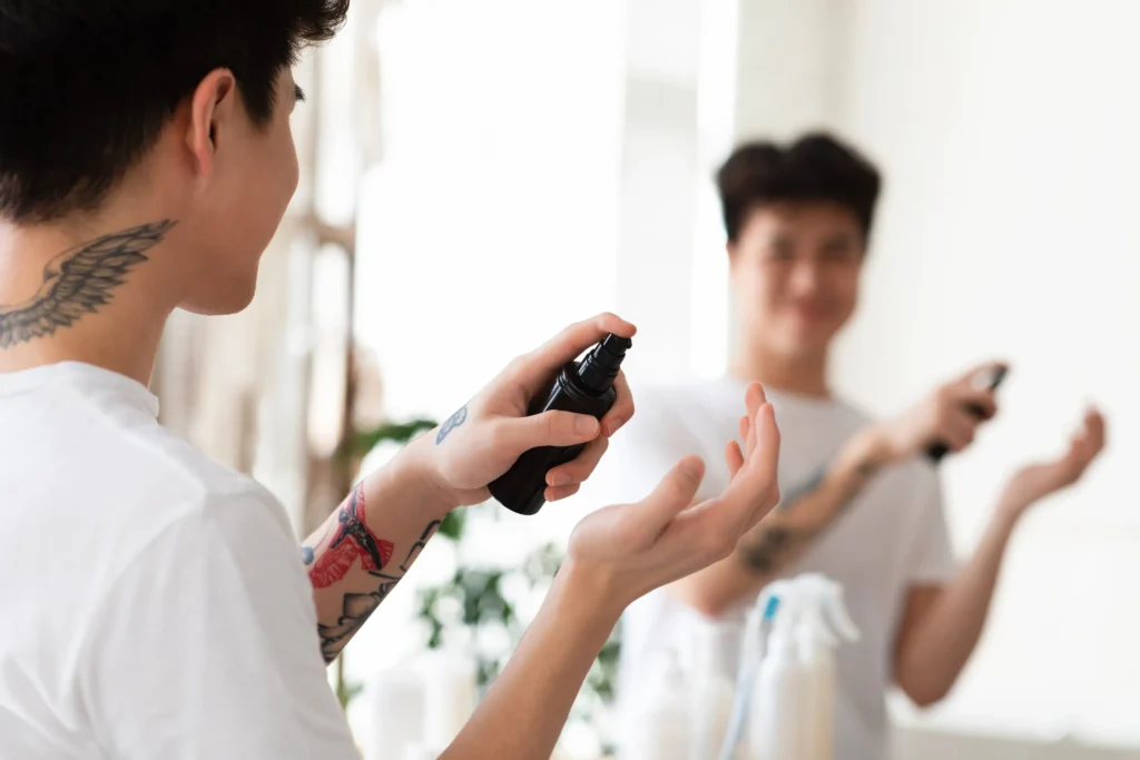 Joven tatuado rociando perfume Valentino hombre en su mano mientras mira su reflejo en el espejo del baño.